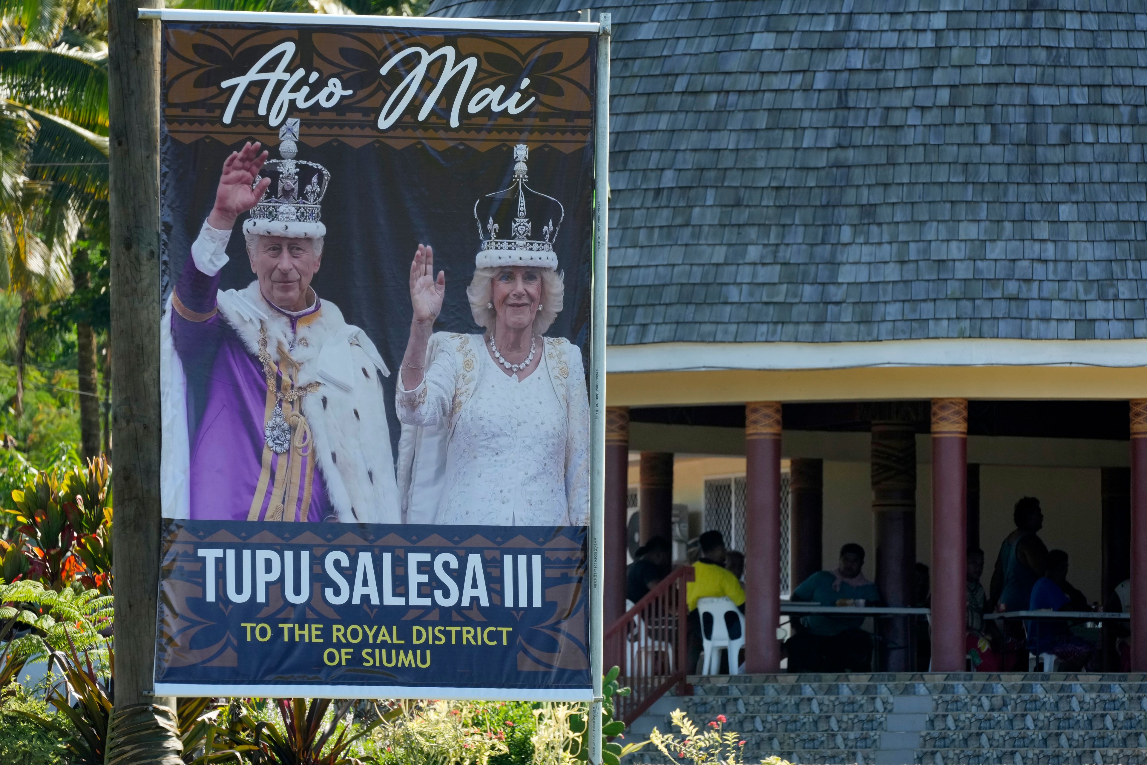 People gather together at a meeting place near a portrait of King Charles III and Queen Camilla in the village of Siumu, Samoa, on Monday, Oct. 21, 2024, as the village prepares for the arrival of the royals. (AP Photo/Rick Rycroft)