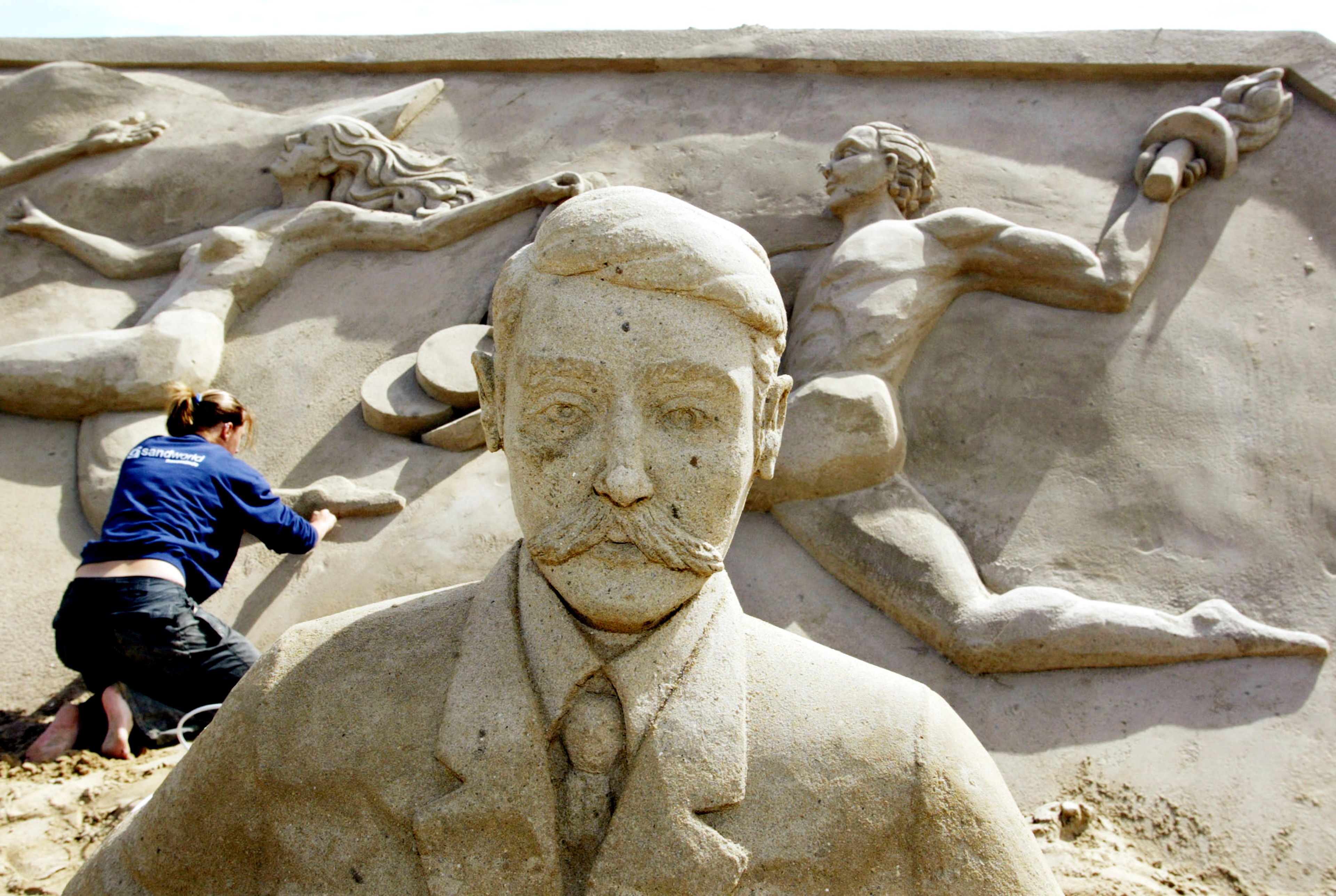 FILE - Carver Maaike Huizinga from the Netherlands gives a final touch to the sand sculpture of sports people behind the sand figure of Pierre de Coubertin, the founder of the modern Olympic Games, in the "Olympic Stadium Travemuende" at the beach of the Luebeck Bay in Luebeck-Travemuende, northern Germany on Tuesday, July 6, 2004. Coubertin envisioned the Olympics as a pacifist exercise that could foster international cooperation and peace, especially in the wake of France's defeat in the Franco-Prussian War. (AP Photo/Heribert Proepper, File)